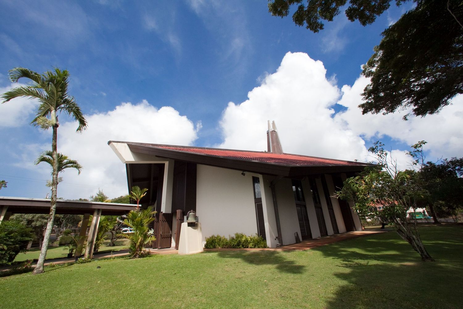 Modern Hawaiian chapel with red roof, palm trees, and lush green lawn under blue sky.