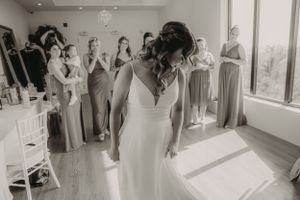 A wedding party getting ready in a sunlit room while bridesmaids in sage green dresses look on with excitement.