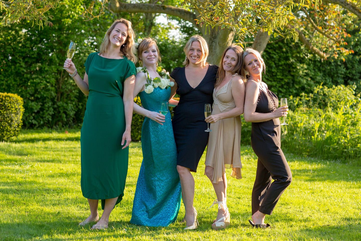 A group of friends in elegant dresses pose together on a sunny lawn.