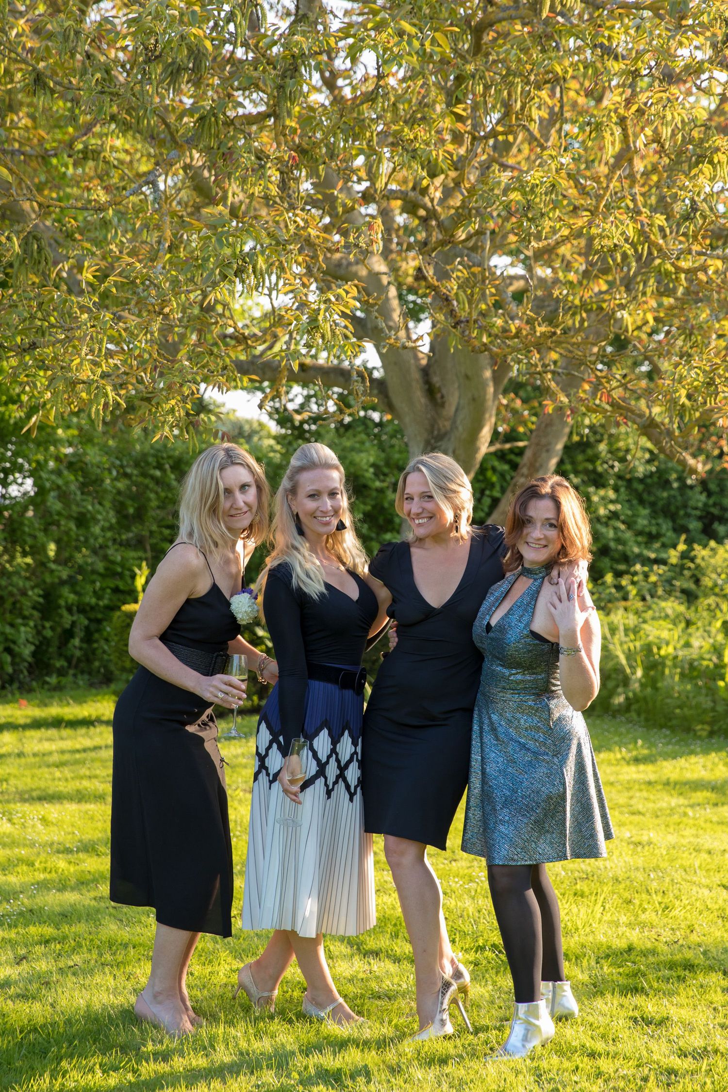 Four friends in stylish dresses pose together on a sunny lawn with trees in the background.