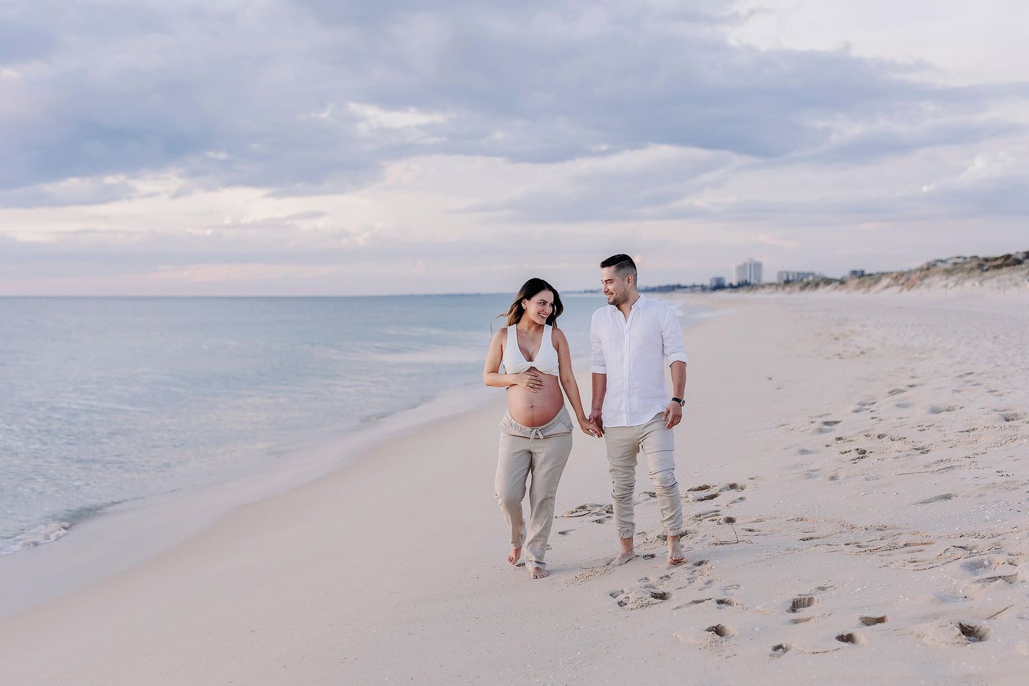 A couple expecting a baby walking along a beach in Perth at sunset, smiling at each other for their pregnancy photoshoot