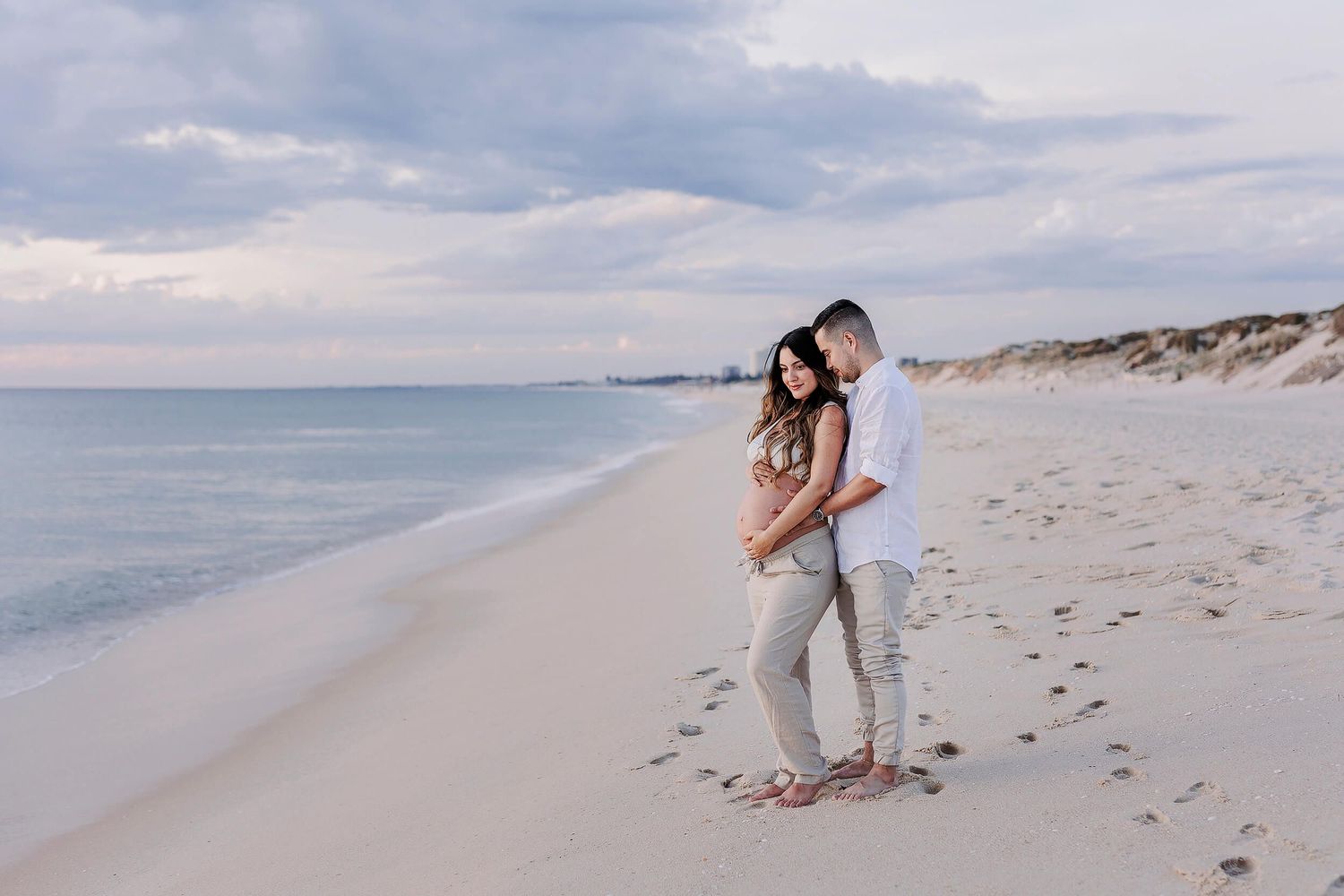 A couple cuddling on the shoreline of the beach in Perth, cradling the woman's pregnant belly for a photoshoot in Perth