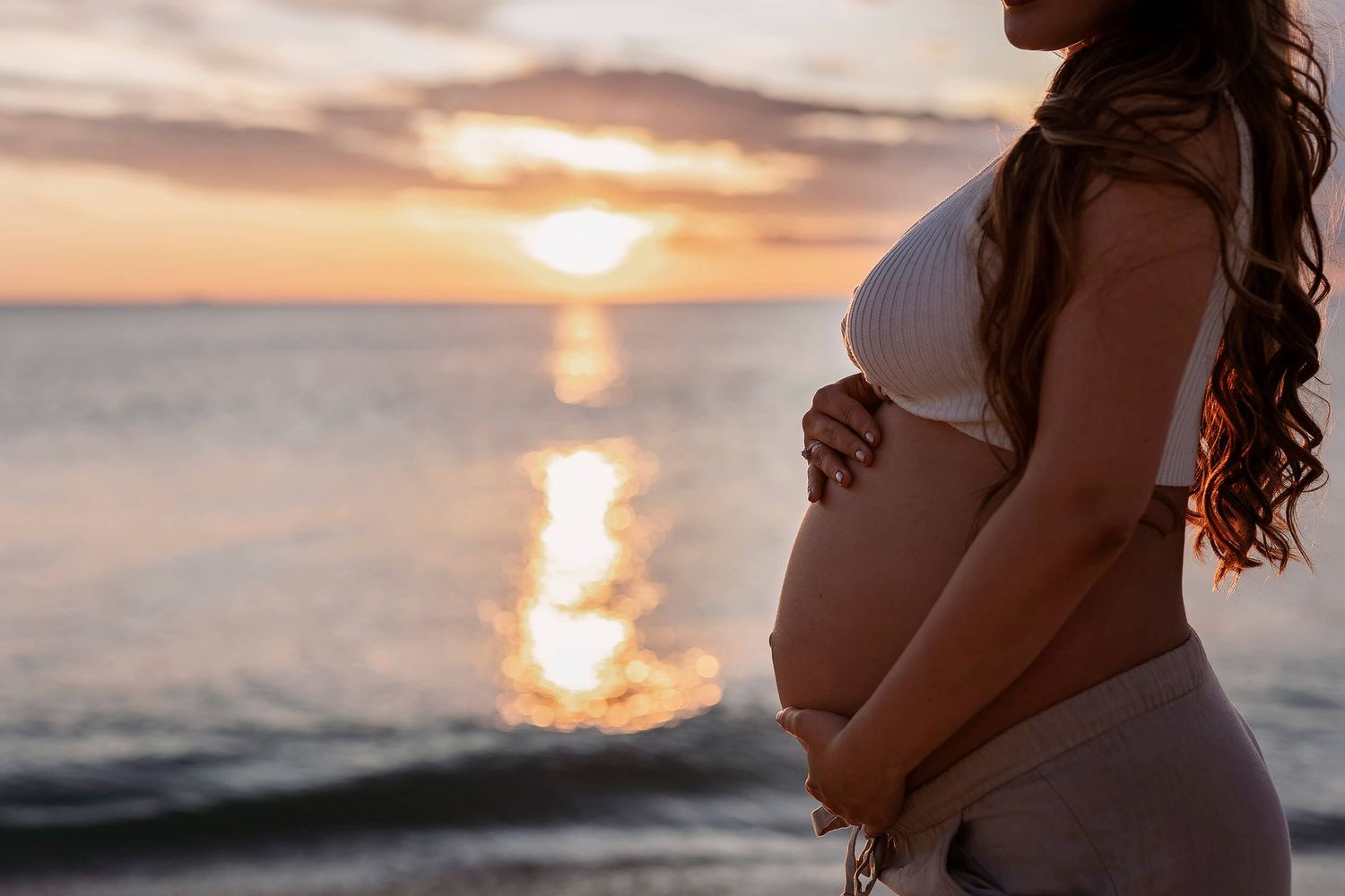 A woman with a naked, pregnant belloy cradling her bump at sunset on the beach during her photoshoot in Perth