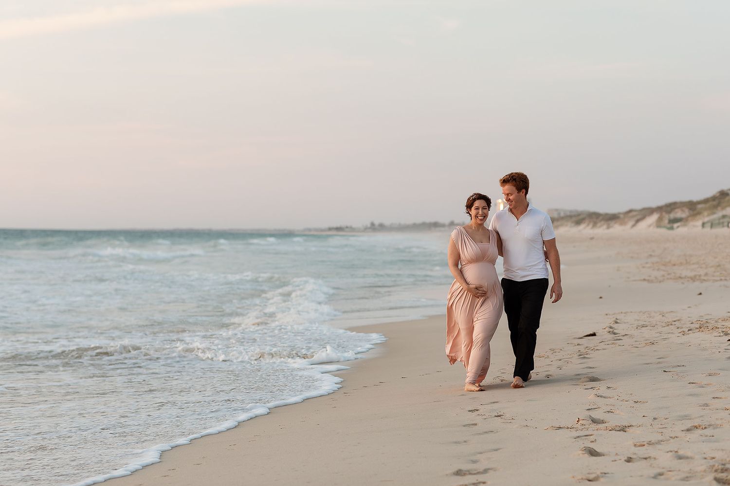 A pregnant woman and her husband walking along the beach and smiling at sunset in Perth for her maternity photoshoot.