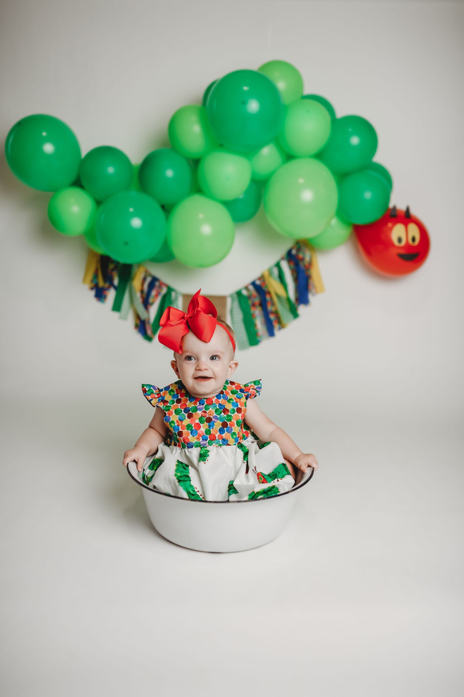 Baby in colorful outfit sitting in metal basin under green balloon arch.