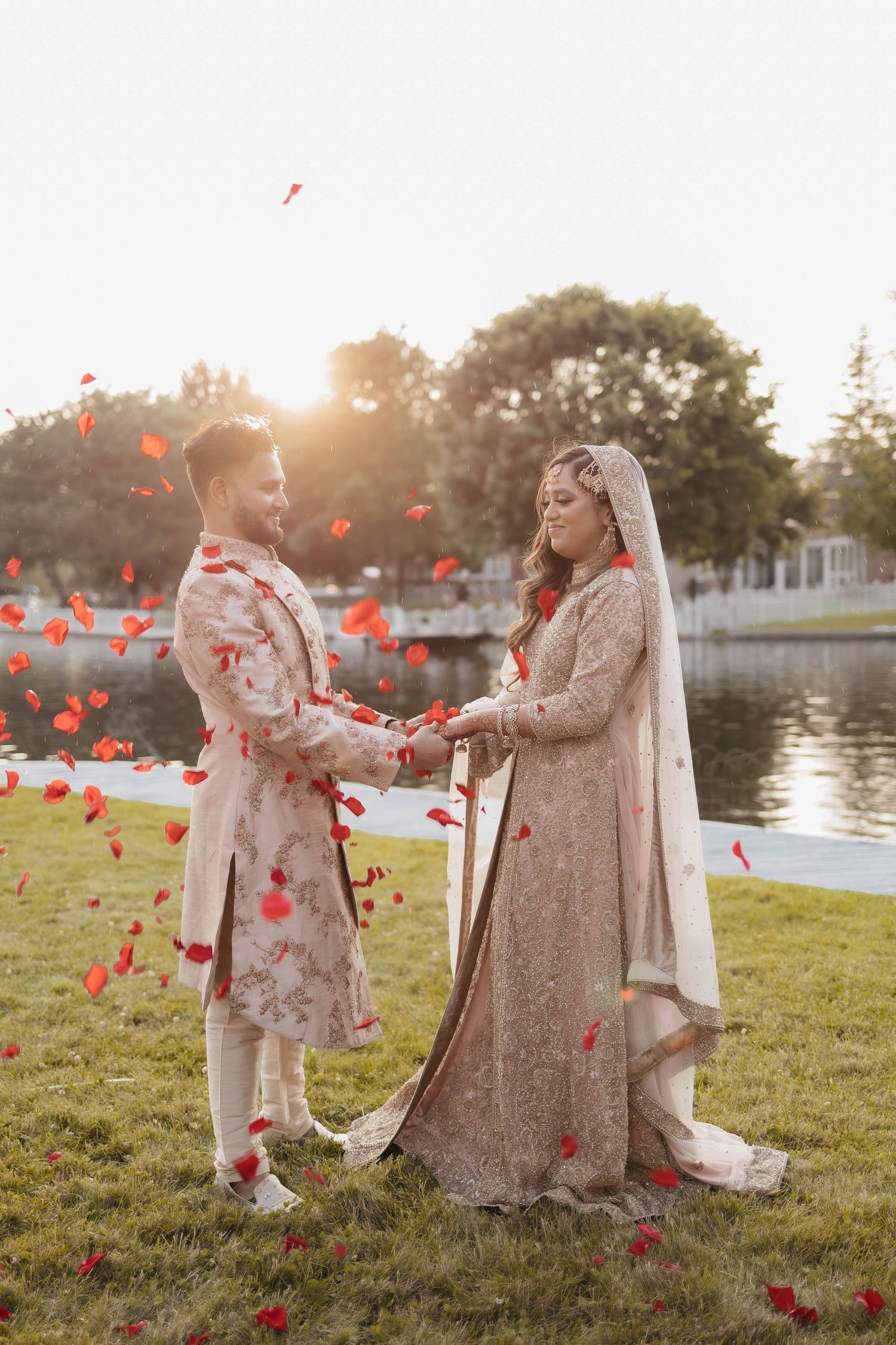 Newlyweds share a romantic moment outdoors with red rose petals falling in the golden sunlight.