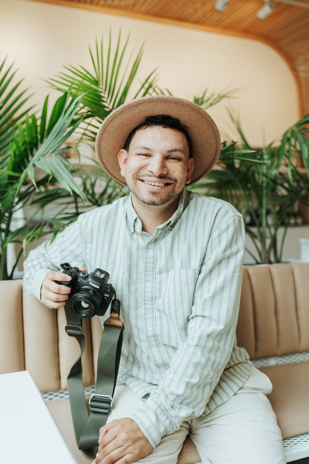 Photographer wearing striped shirt and hat sits on yellow couch with camera in tropical setting.