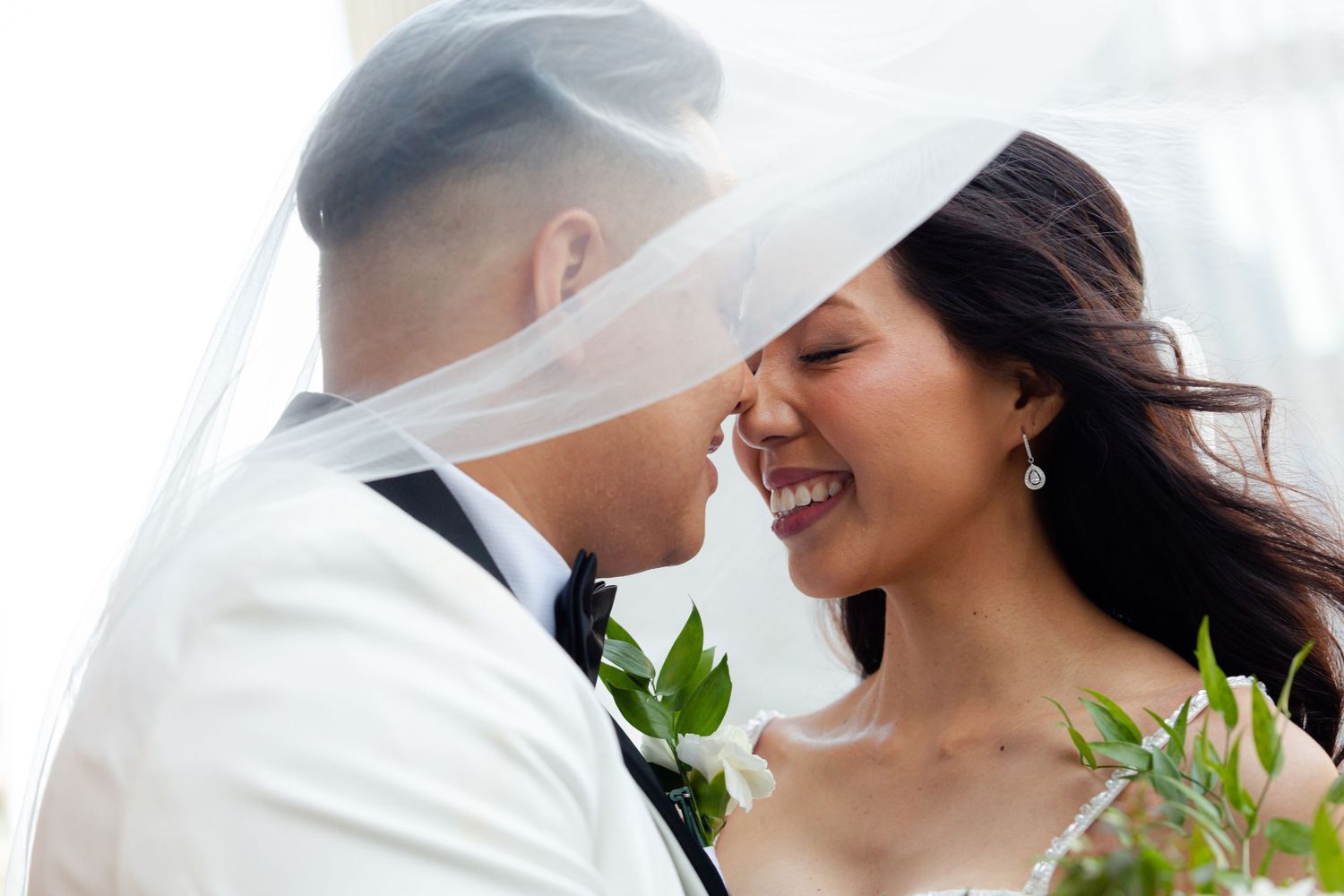 Romantic wedding couple share intimate moment under flowing veil outdoors.