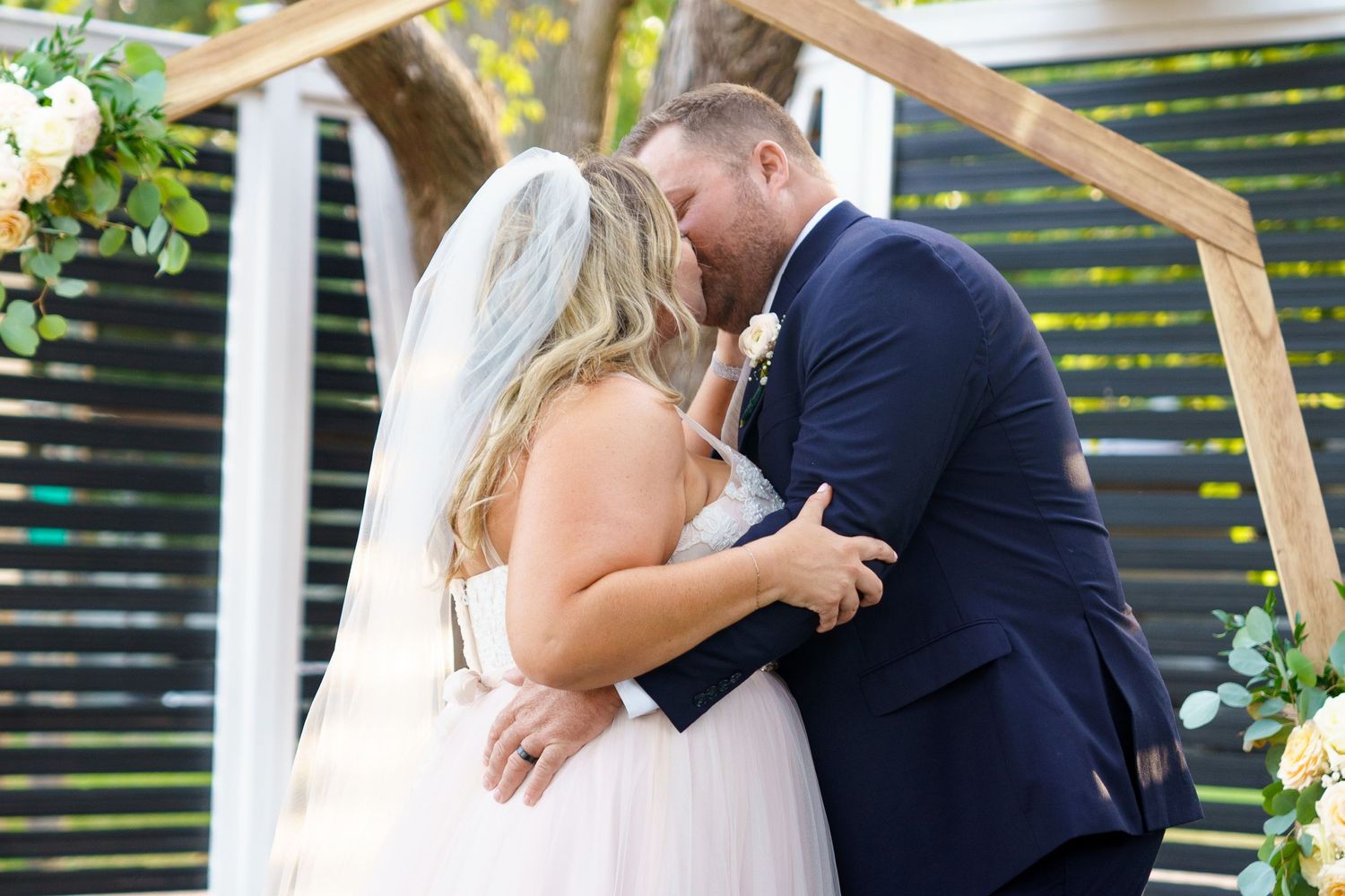 Intimate moment between newlyweds under wooden arch decorated with flowers.