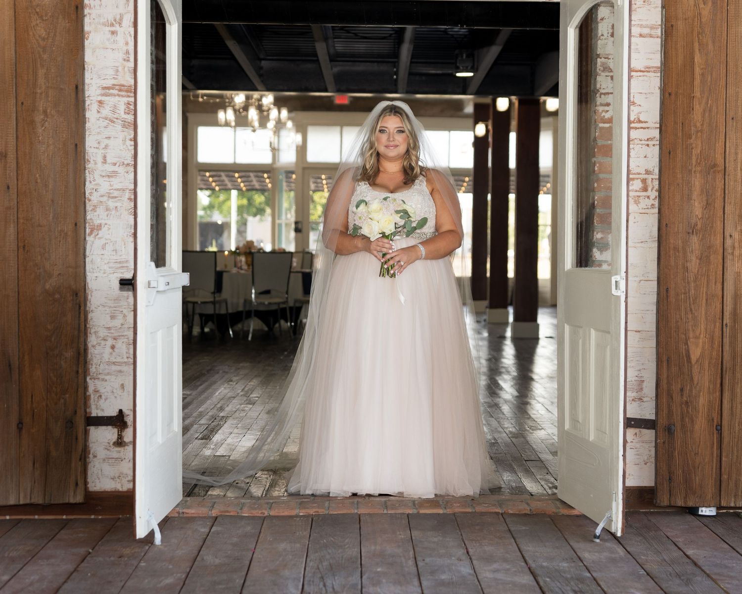 Bride in flowing white tulle gown stands in rustic wooden doorway holding bouquet.
