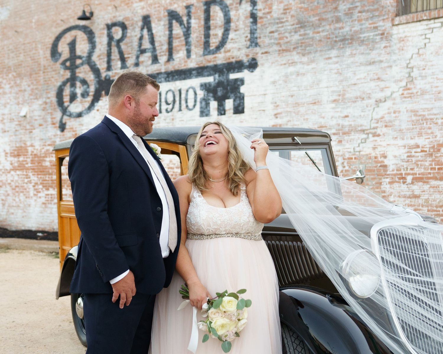 Happy couple laughs together in front of vintage car and rustic brick wall.