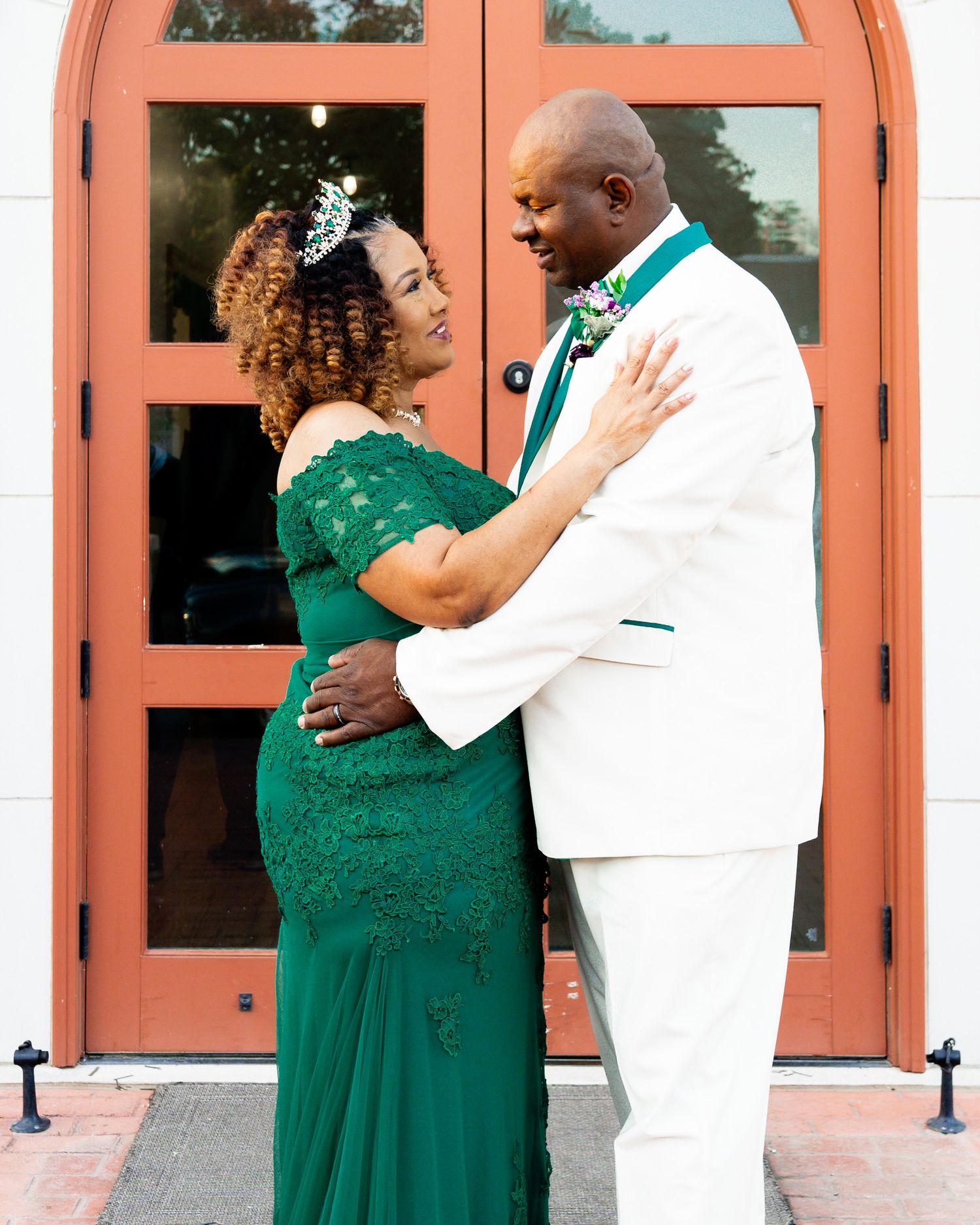 Couple in emerald green dress and white suit embrace romantically in front of coral-colored doors.