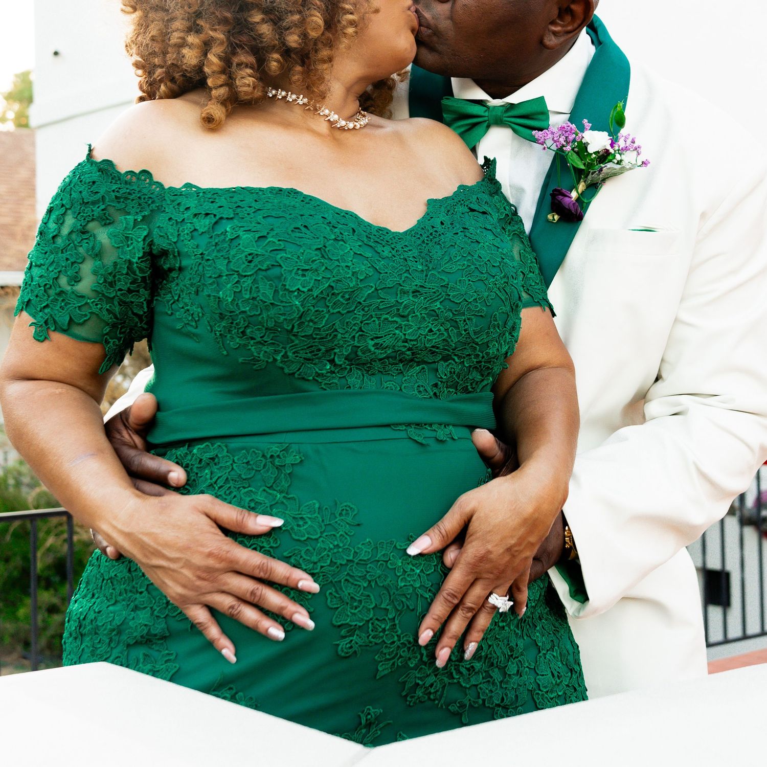 Close-up of couple's embrace showing intricate lace details of emerald green dress against white suit jacket.
