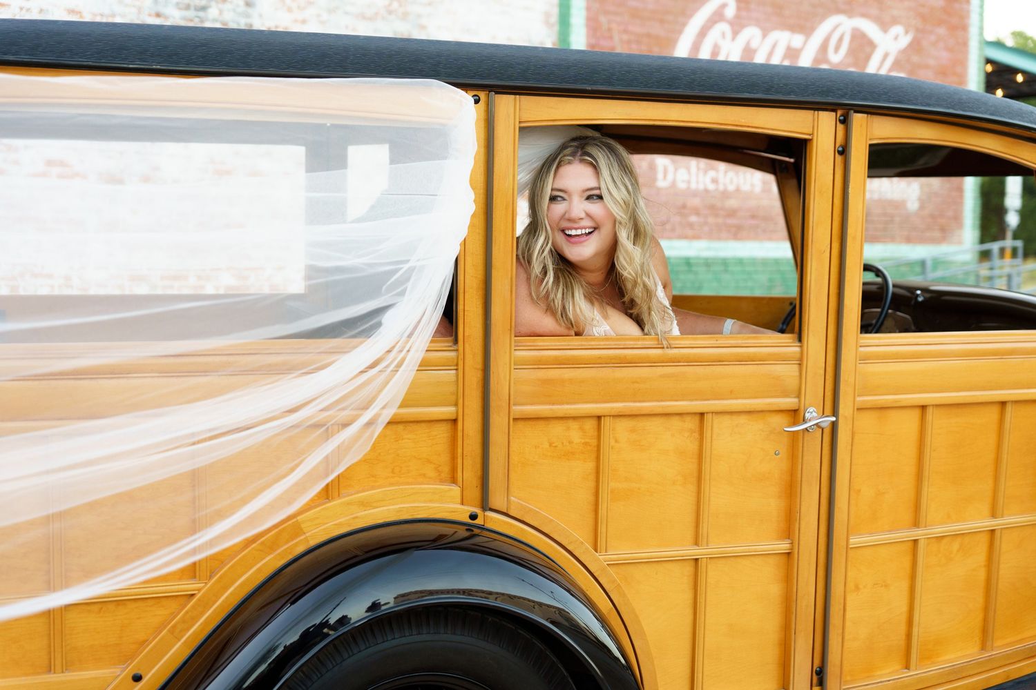 Joyful bride in flowing veil smiles from vintage yellow wooden car.