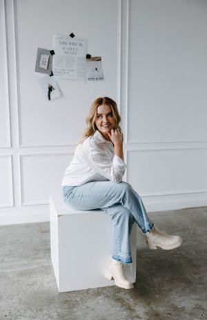 Woman wearing white shirt and light jeans sits casually on white cube in minimalist studio setting.