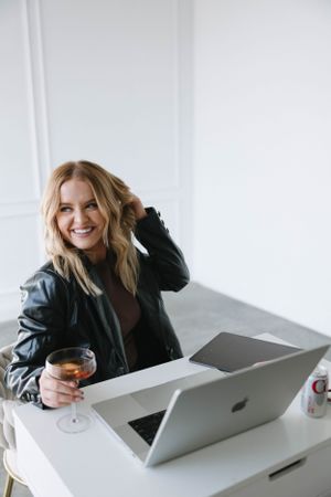 Woman in leather jacket works on laptop with drink at minimalist white desk.