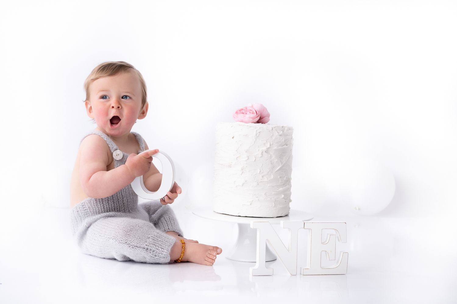 Baby in grey outfit sitting beside white cake on stand.