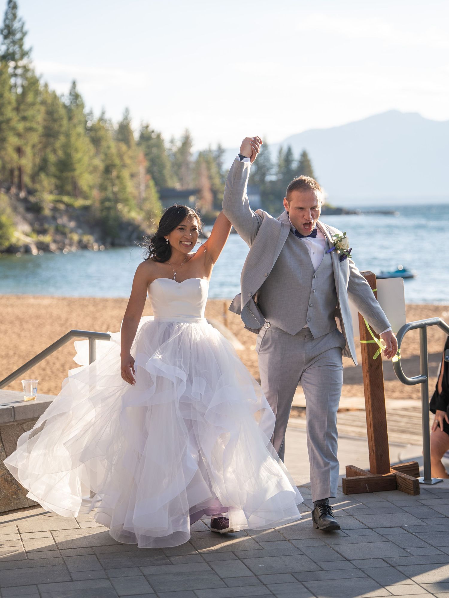 A joyful newlywed couple celebrates on a scenic deck overlooking a lake with mountains and pine trees in the background.