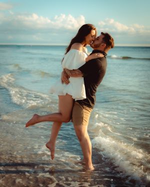 Romantic couple sharing an intimate moment on a sandy beach with waves and blue sky in the background.