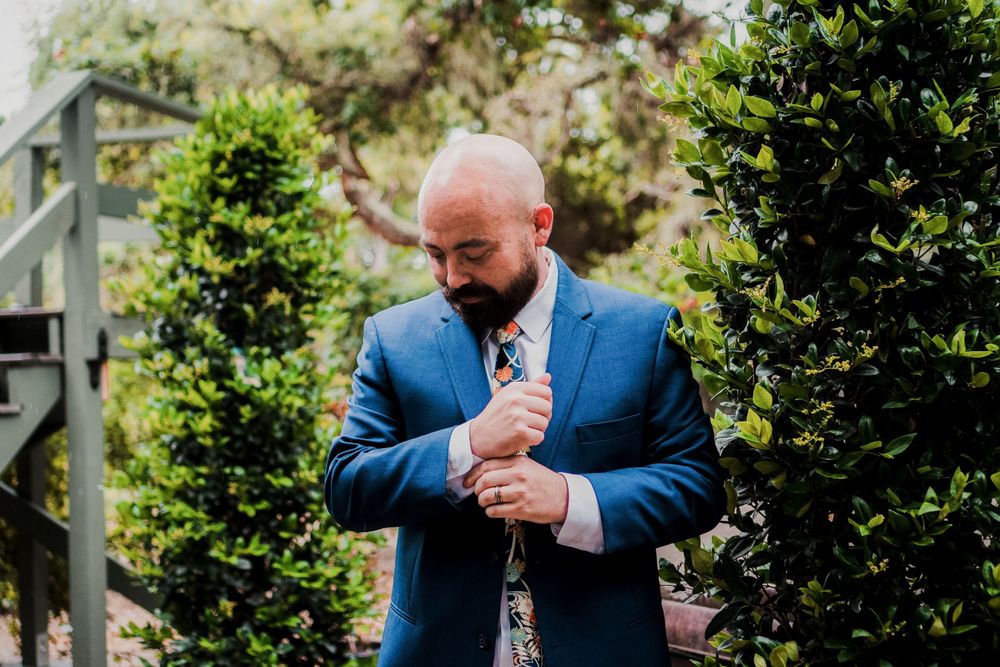 A groom adjusts his tie while wearing a fitted blue suit in a garden setting.