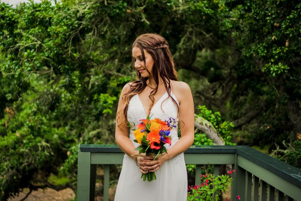 A bride holds a colorful bouquet while standing on a wooden deck surrounded by greenery.