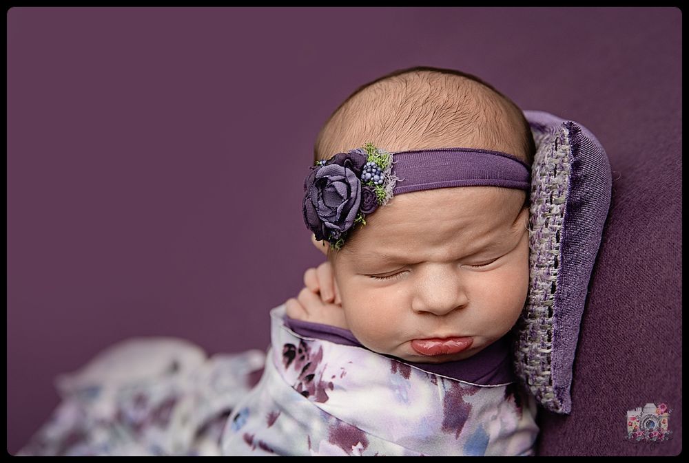 Sleeping newborn wearing purple floral headband against purple background.