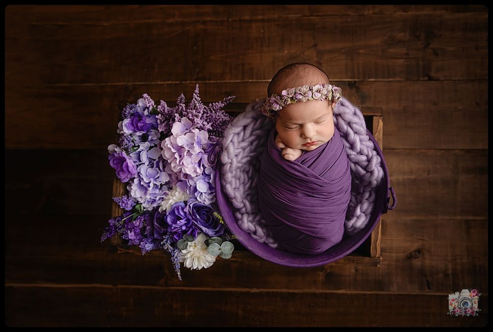 Newborn wrapped in purple swaddle with purple flowers on wooden surface.