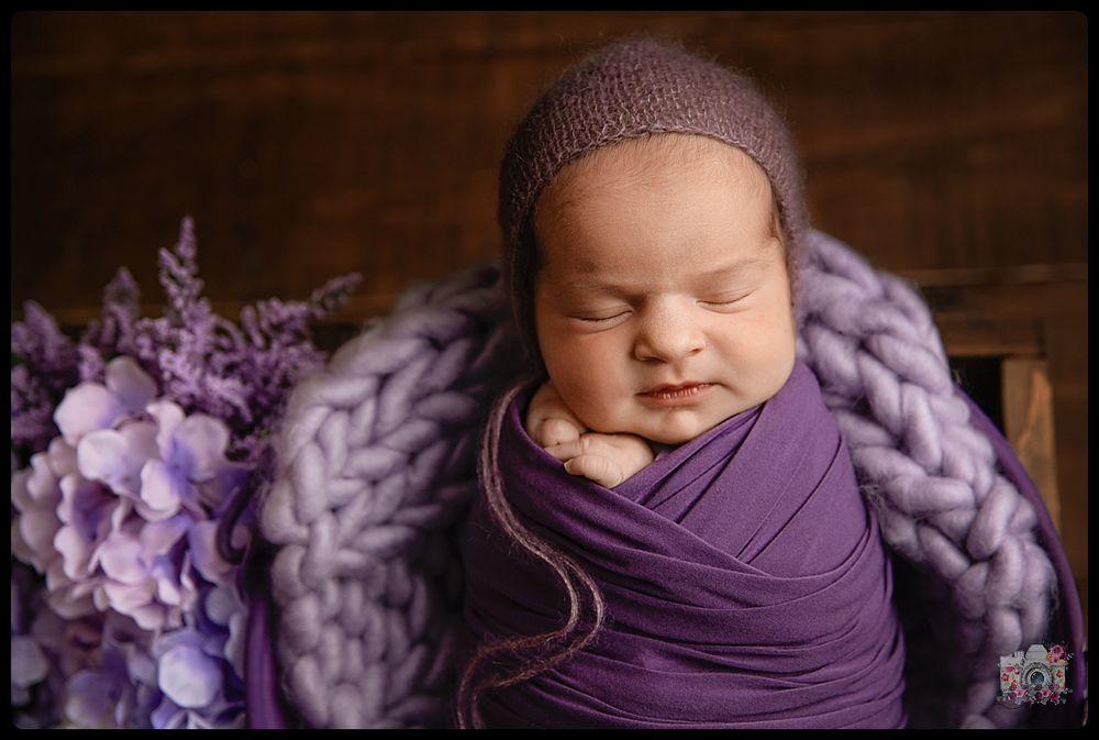 Sleeping baby wrapped in purple swaddle with chunky knit blanket and flowers.