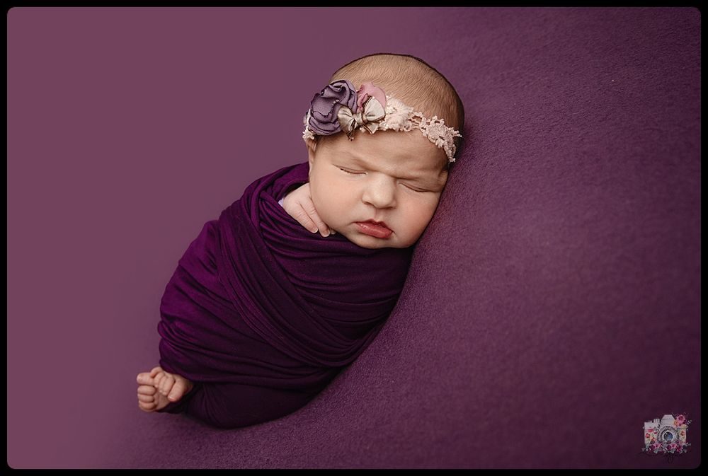Sleeping newborn wrapped in purple fabric wearing delicate floral headband against purple backdrop.