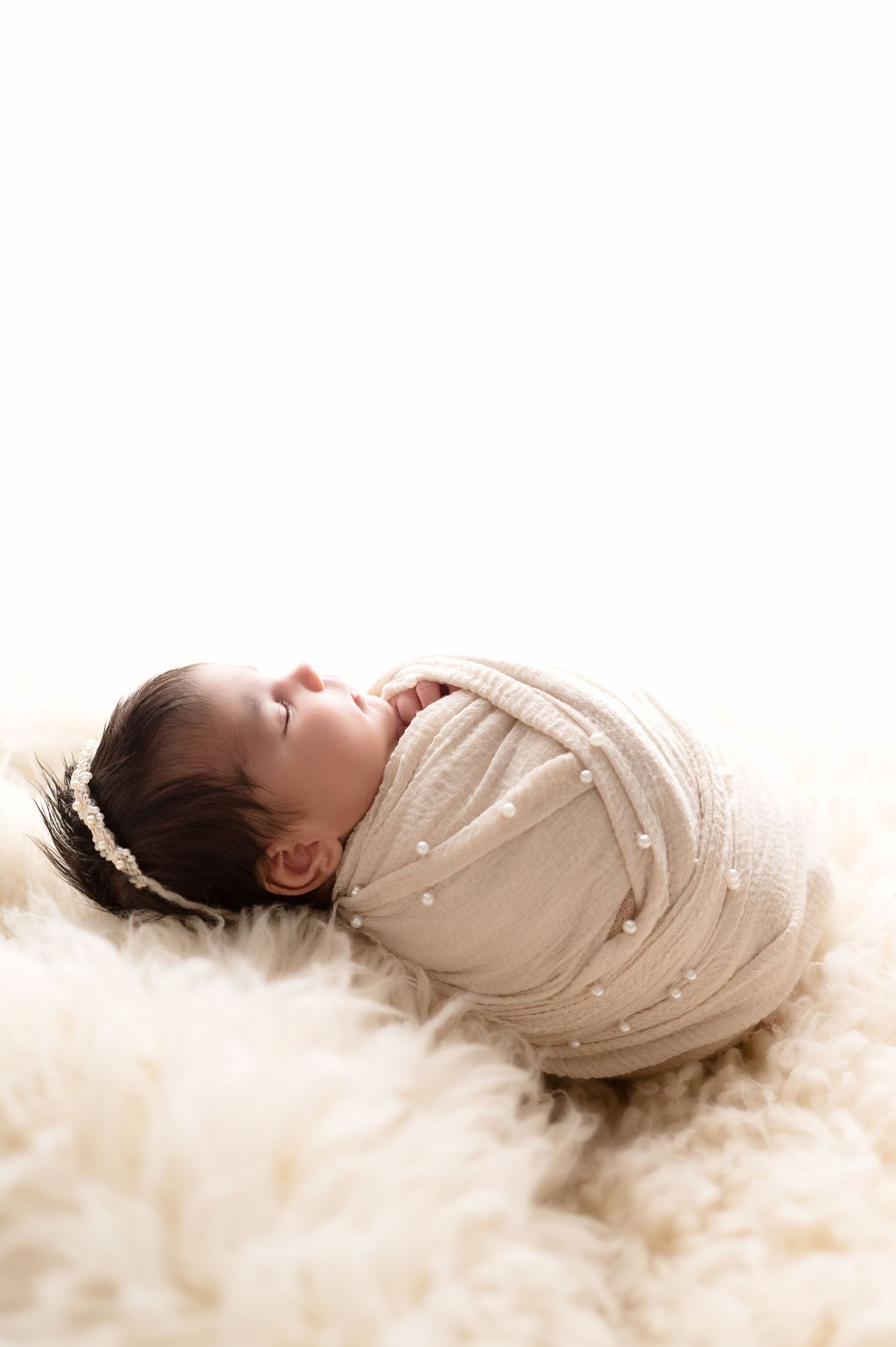 A swaddled newborn sleeps peacefully on creamy white fur background.