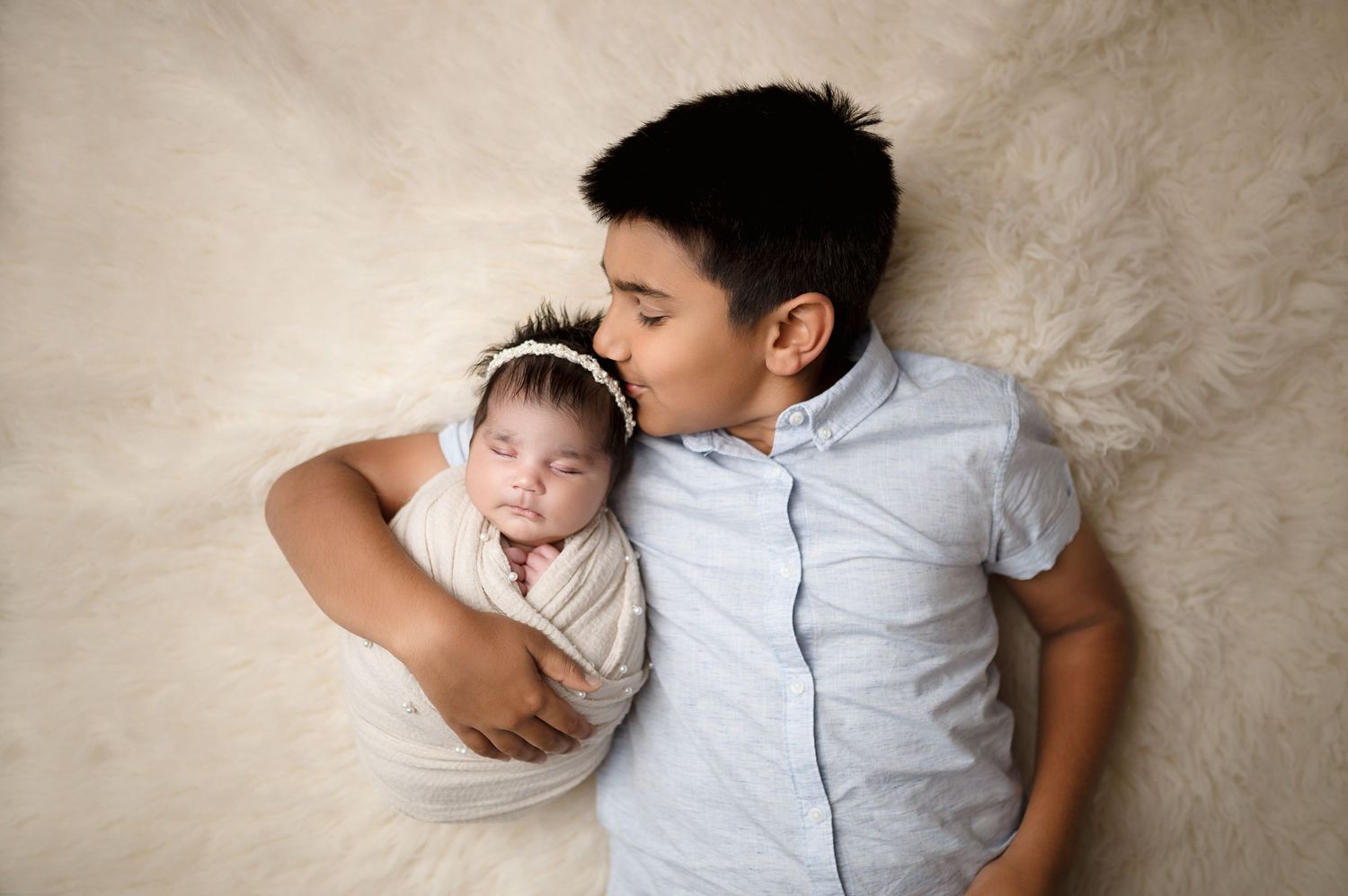 A tender portrait of a person lying with sleeping newborn on cream-colored backdrop.