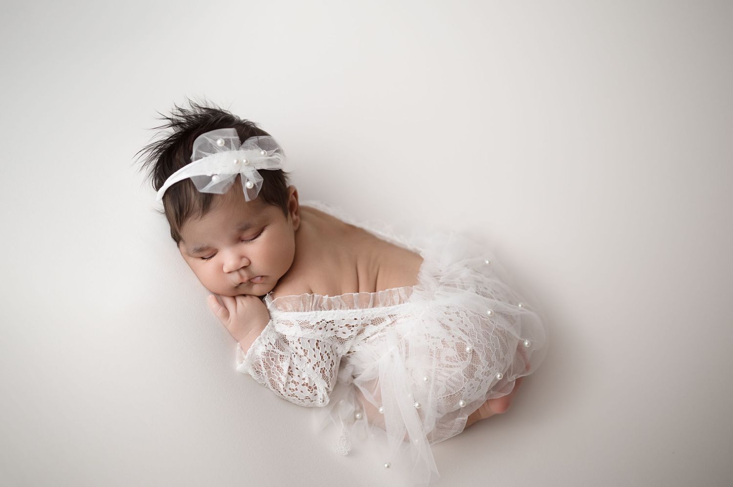 A sleeping newborn wrapped in delicate white lace and headband poses on a neutral background.