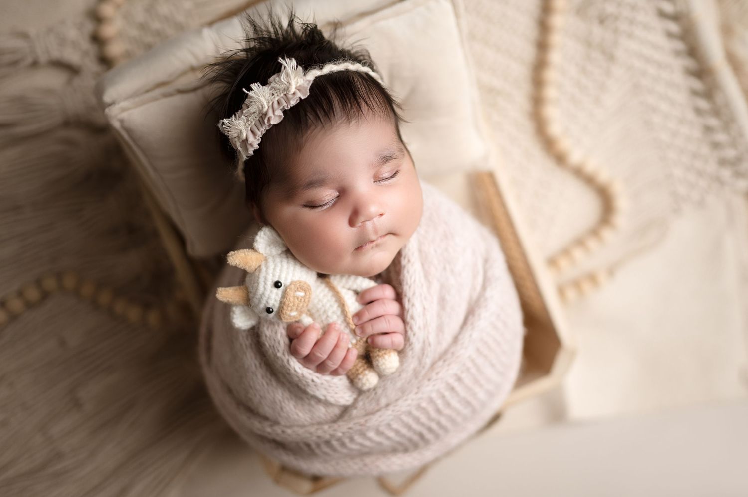 A sleeping newborn in knit wrap holds tiny teddy bear while resting on beige chair.
