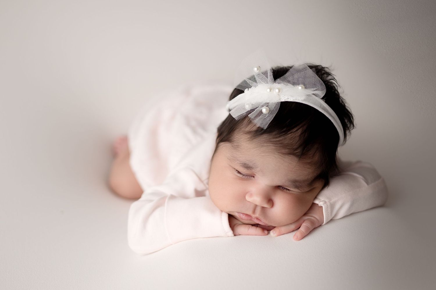 Sleeping newborn wearing pink outfit and white headband rests peacefully on white backdrop.