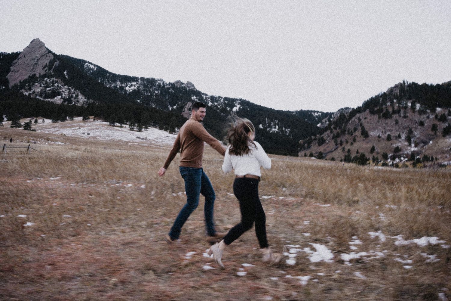 Couple running and laughing together in a snowy mountain meadow during a winter engagement session.