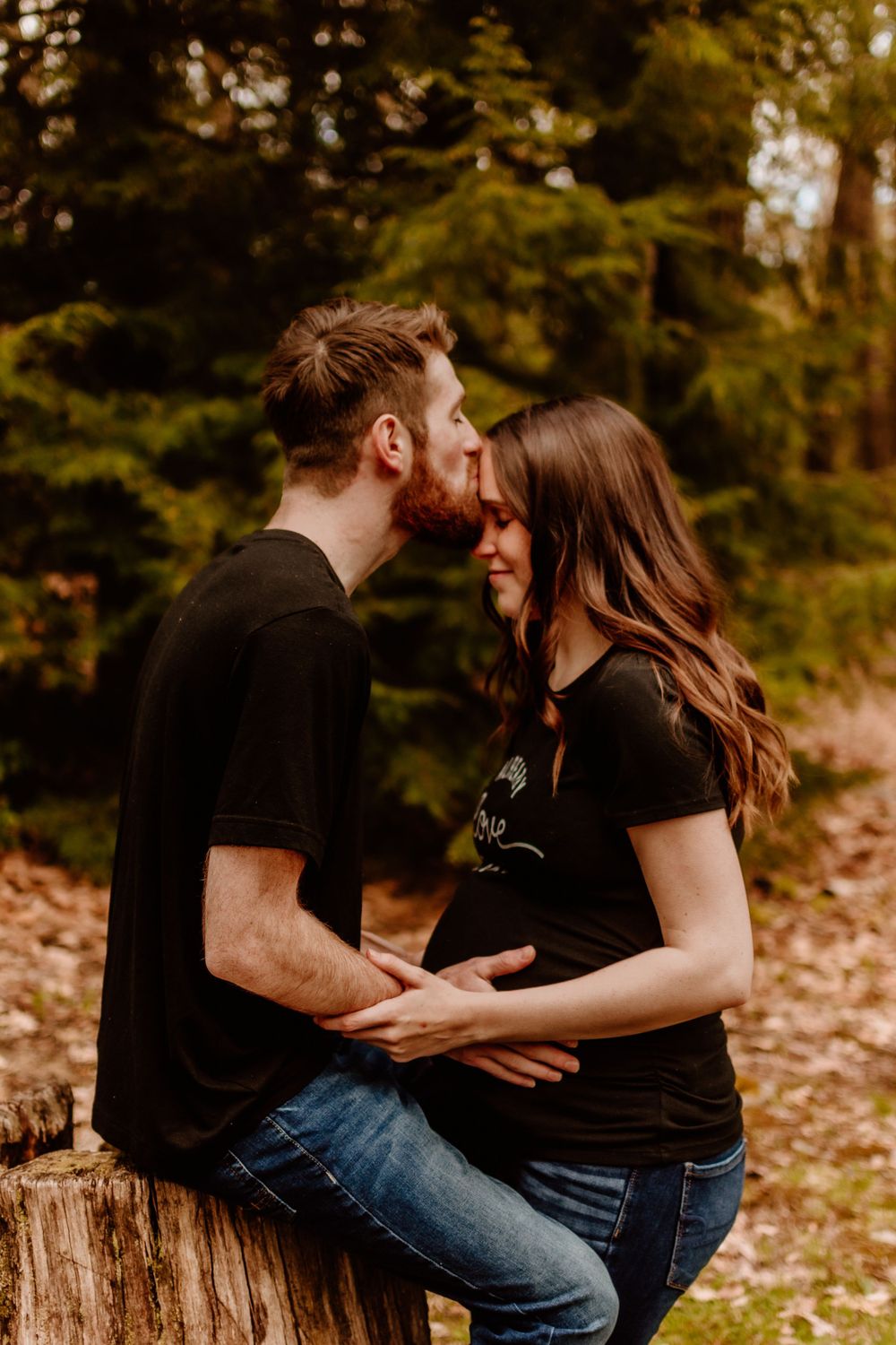 Romantic couple in matching black outfits sharing intimate moments on a tree stump in a forest setting.