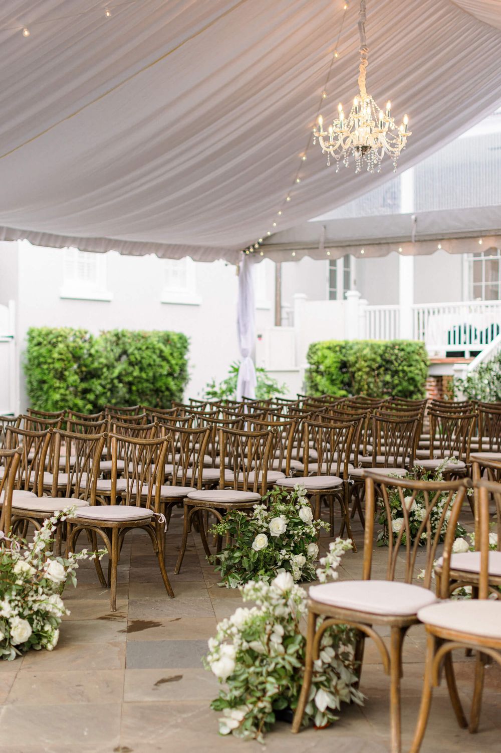 Elegant wedding ceremony setup under a white tent with wooden chairs and lush floral aisle decorations.