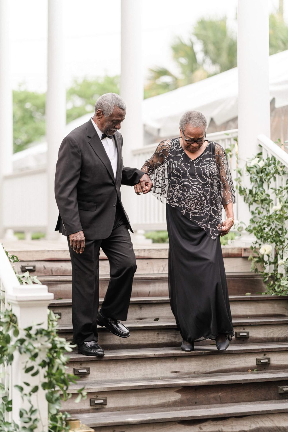 Two elegantly dressed figures carefully descending outdoor stairs at a formal event.
