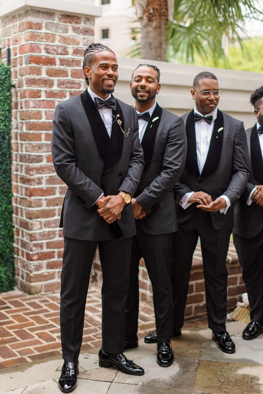 Groomsmen in black tuxedos smiling and standing near a brick wall.