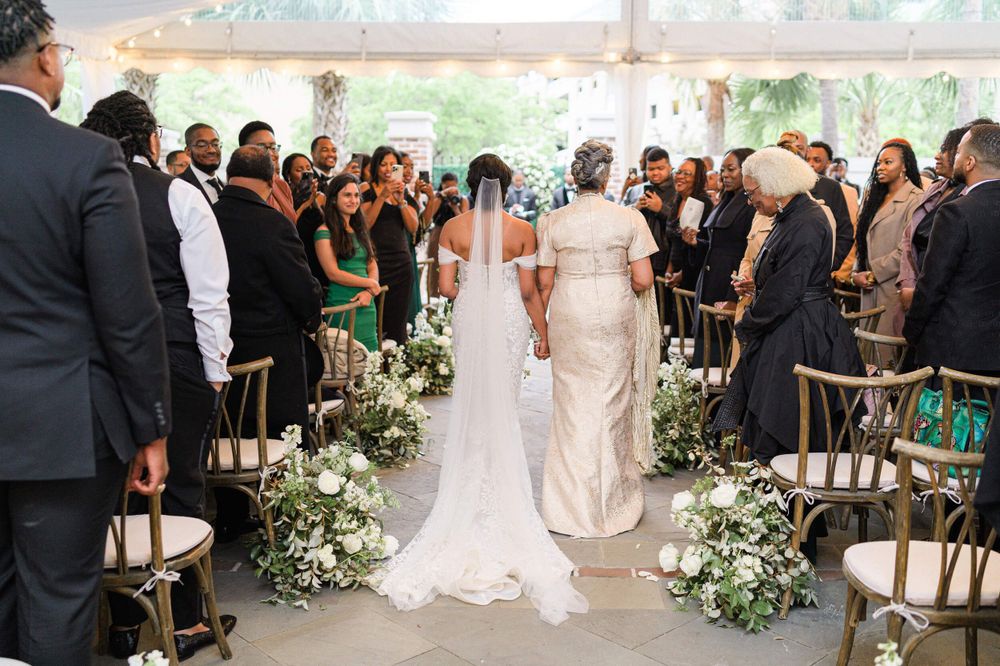 Wedding ceremony processional with white floral aisle decorations and cross-back chairs.