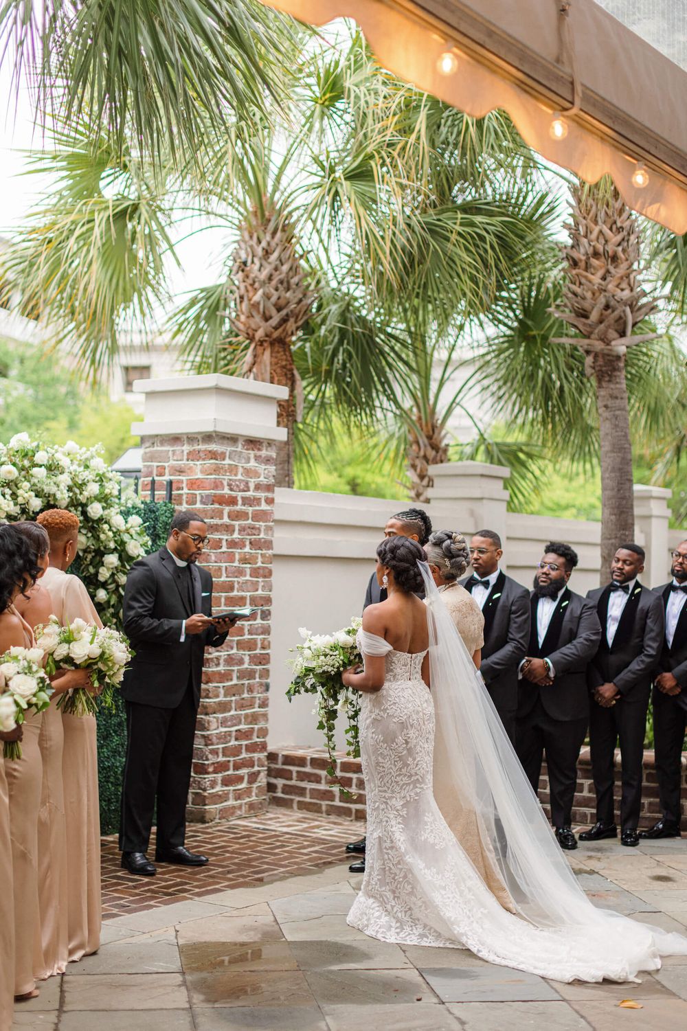 Outdoor wedding ceremony under palm trees with formal wedding party and floral arch.