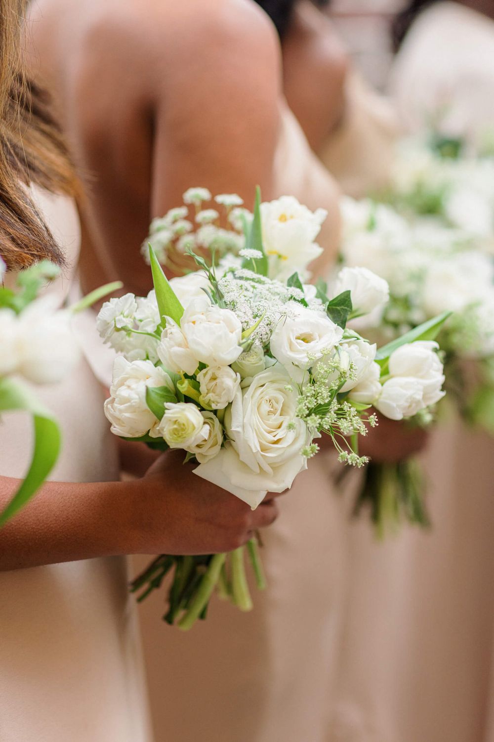 White wedding bouquet with roses and delicate greenery held by hands against a soft blush background.