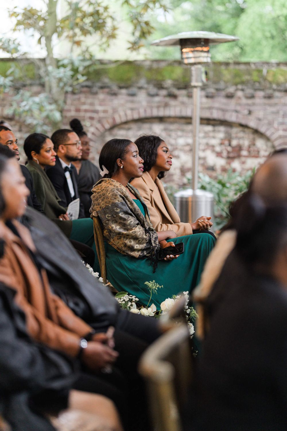 Wedding guests react emotionally during an outdoor ceremony with brick and greenery backdrop.