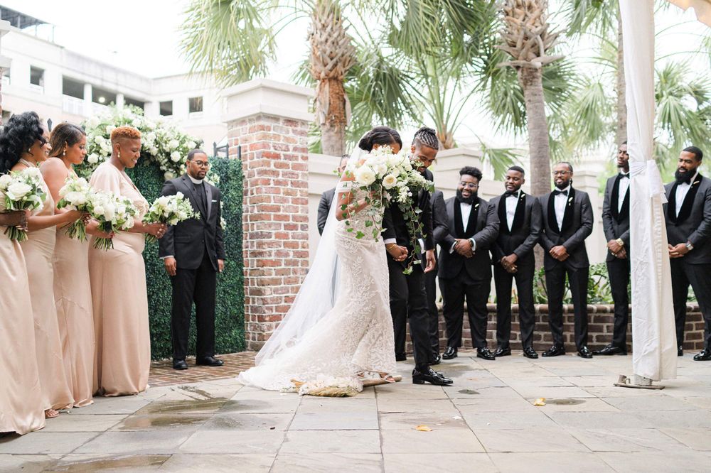 Wedding party stands in formation during outdoor ceremony with palm trees and brick columns.