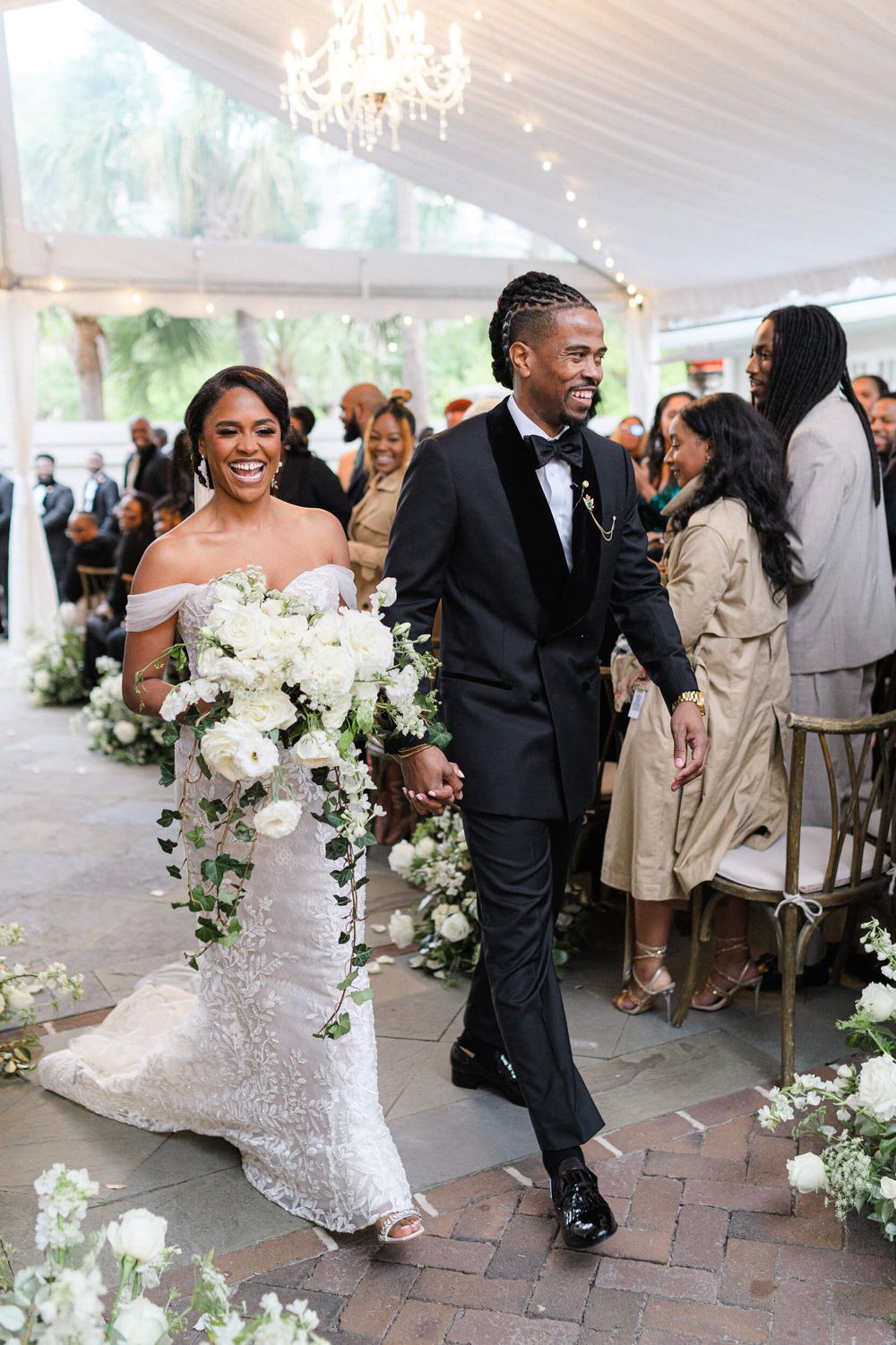 Newlyweds walk down aisle under elegant white tent decorated with chandeliers and flowers.