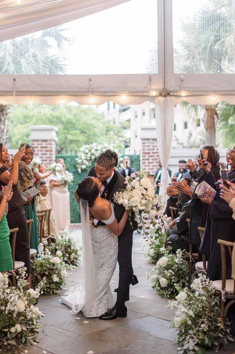 Wedding ceremony kiss under tent with floral aisle decorations and guests watching.