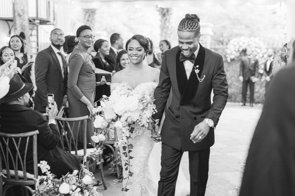 Black and white photo of newlyweds walking down aisle with guests seated on both sides.