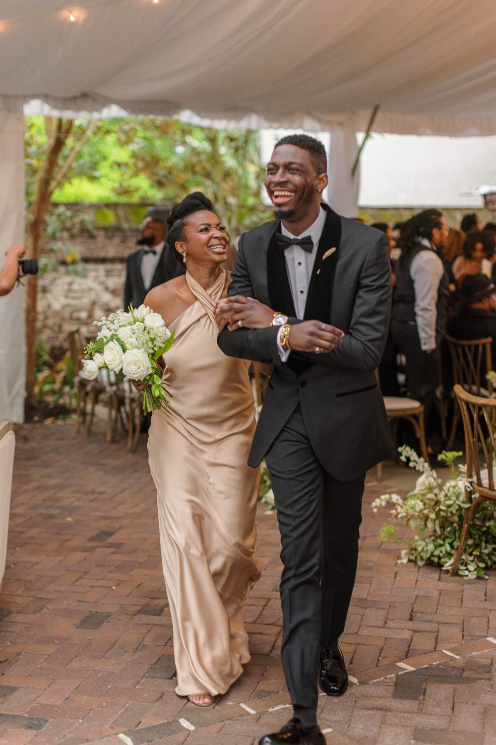 Wedding celebration with joyful couple walking together in formal attire under a tented venue.