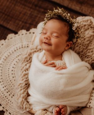 Sleeping newborn wrapped in white swaddle wearing delicate crown on textured blanket.