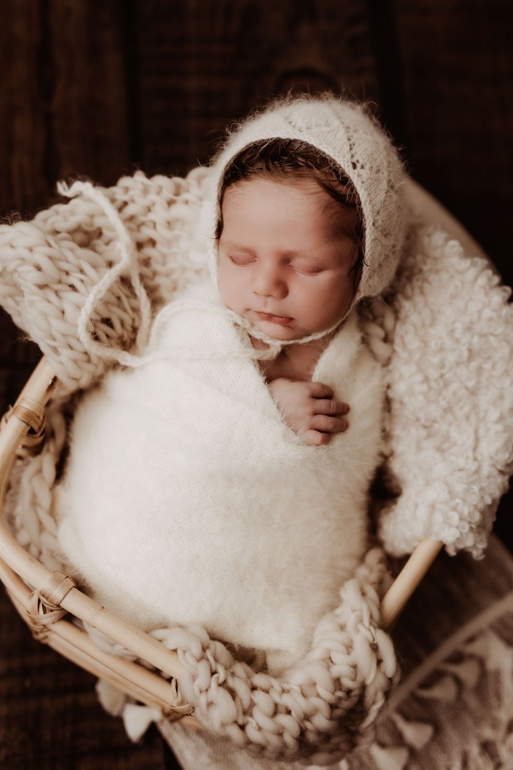 Sleeping newborn in a white bonnet and wrap nestled in a cozy basket.